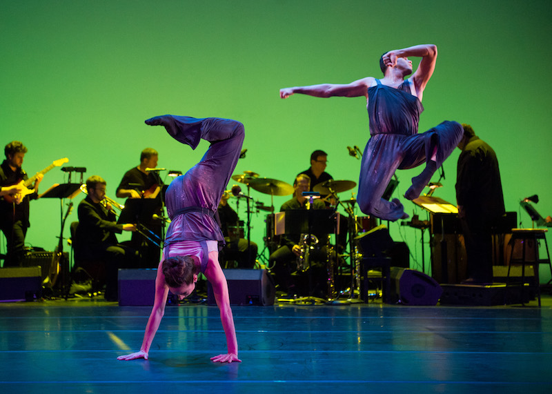 One dancers stands on her hands with her legs bent and splayed in the air. Another dancer jumps high into the air with his feet tucked underneath him.